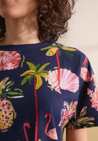 Chemise à manches courtes marine avec des imprimés tropicaux vibrants, présentant des fleurs roses, des feuilles vertes et des flamants roses. Tissu en coton doux.