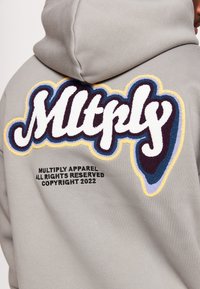 Sweat à capuche gris avec un logo brodé texturé multicolore "Mltply" au dos, avec un texte noir en dessous indiquant les informations de copyright.