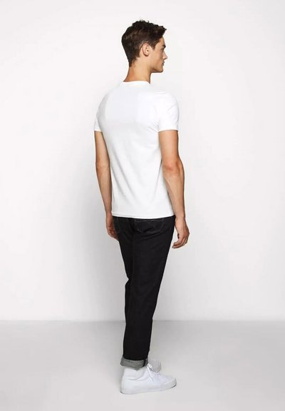 T-shirt bianca a maniche corte con una texture liscia, abbinata a jeans scuri e sneakers bianche, vista da dietro.