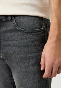 Vaqueros de mezclilla negros con un corte ajustado, que cuentan con un bolsillo frontal, un ligero desgastado y una textura suave, complementados por una camisa de color claro.