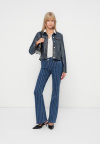 Denimjakke med sølvknapper, brukt over en hvit strikket topp, kombinert med høytsittende stripete brystbukser og svarte høyhælte sko.