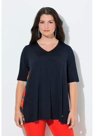 Kvinde med langt mørkt hår iført navyblå V-hals top med farverige sidepaneler og lyse røde bukser, stående op ad en hvid væg.