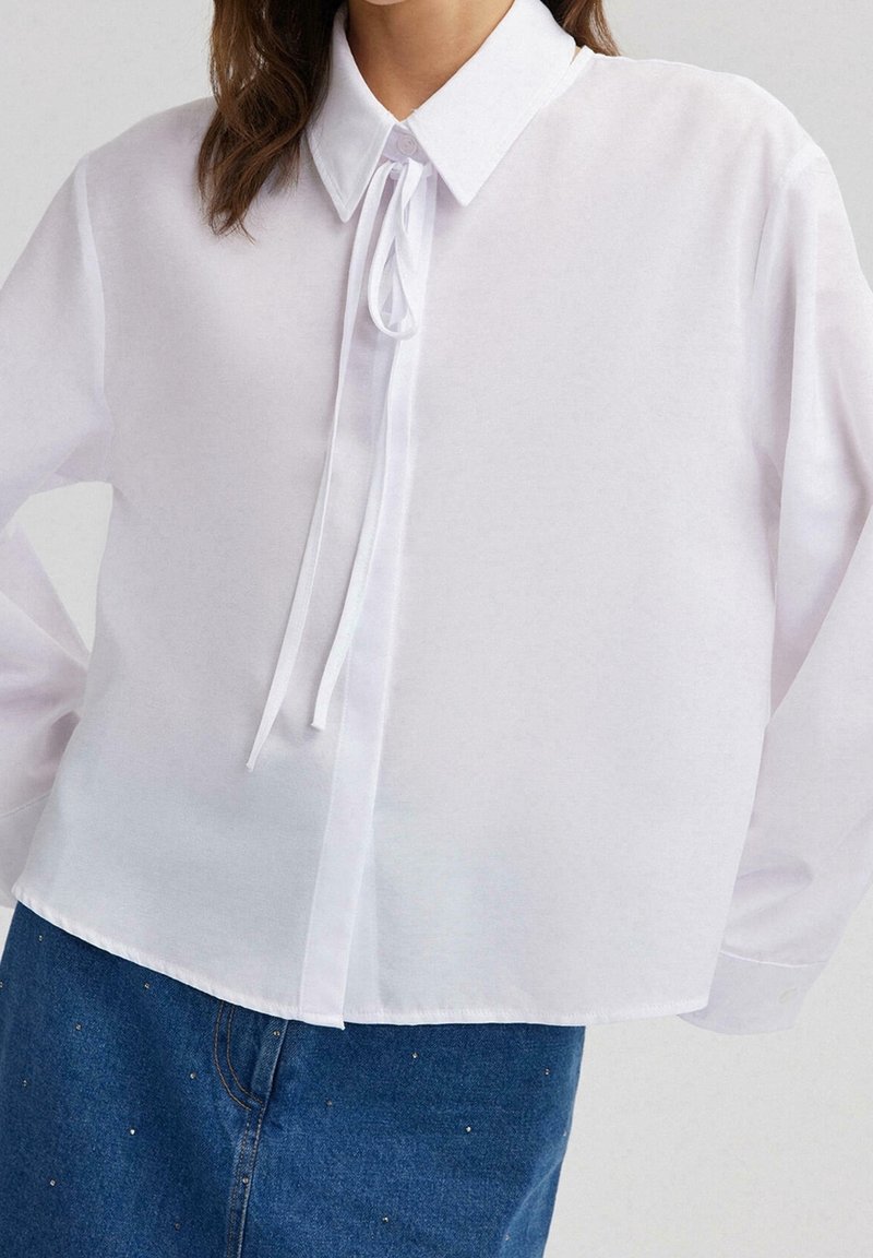 Blouse blanche à manches longues avec col pointu et détail de nouage à l'avant, associée à un jean en denim bleu avec de petites décorations cloutées.