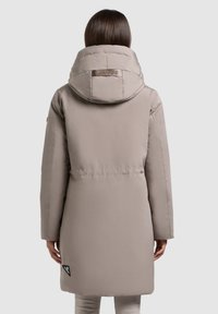 Lys beige pufferjakke med en stor hætte og et logo-mærke nær kanten. Har en glat tekstur og et mellemlangt design.