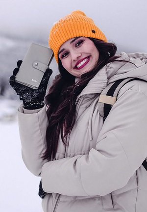 Gray wallet with a flap and logo held against a snowy background. The person wears a beige puffer coat and an orange knitted beanie.