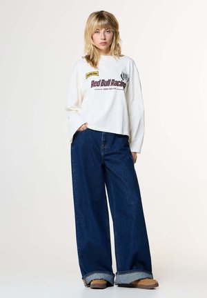 Junge Frau mit blondem Haar, die einen weißen Red Bull Racing Pullover, weit geschnittene dunkelblaue Jeans und braune Schuhe trägt, steht vor einem hellen Hintergrund.