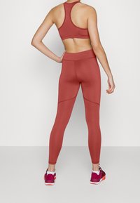 Set di reggiseno sportivo e leggings color marrone, con texture a costine, vita alta e design aderente. Le scarpe sono di colore bordeaux con accenti neri.