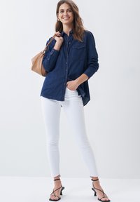Camisa de mezclilla en azul oscuro con botones en la parte delantera, dos bolsillos en el pecho y mangas largas. Combinada con jeans blancos ajustados y tacones negros con tiras.