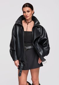 Vrouw draagt een oversized zwarte leren jas, zwarte strapless top, zwarte minirok met riem en zwarte choker, staand tegen een effen achtergrond.