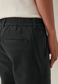 Schwarze Hose mit elastischem Bund, ausgestattet mit einer einzigen Gesäßtasche und einem seitlichen Reißverschluss. Der Stoff wirkt weich mit einer matten Textur.