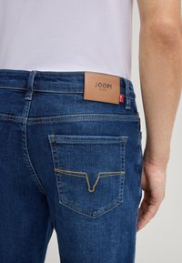 Denim jeans in dark blue with a leather label reading "JOOP!" at the back. Features yellow stitching on the pockets and a slim fit design.