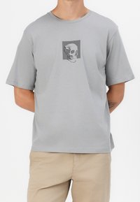 Grå bomulls-T-shirt med korta ärmar, med en central grafisk bild av ett skalle. Avslappnad passform, lätt tyg och enkel rund halsringning.