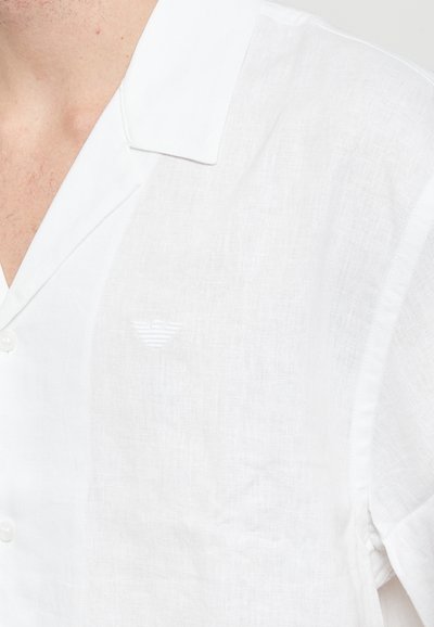 Camicia bianca in lino a maniche corte con una consistenza morbida, caratterizzata da un logo ricamato discreto vicino al petto e chiusura con bottoni.
