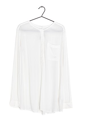 Camicia bianca a maniche lunghe con colletto rotondo, chiusura con bottoni, una tasca sul petto e un tessuto morbido e trasparente, appesa a un gancio nero.