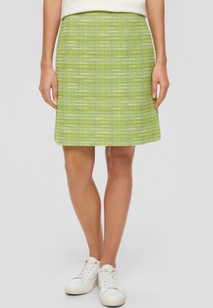 Kobieta w zielonej, teksturowanej spódnicy w kształcie litery A i białych tenisówkach, stojąca na tle gładkiego, jasnoszarego tła.