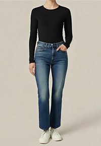 Personne portant un haut noir à manches longues, un jean bleu taille haute légèrement délavé, et des chaussures blanches, debout devant un fond uni.