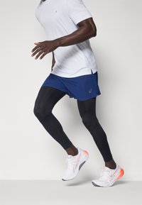 Mannelijke sportoutfit met een witte T-shirt met korte mouwen, blauwe shorts, zwarte leggings en witte hardloopschoenen met oranje accenten.