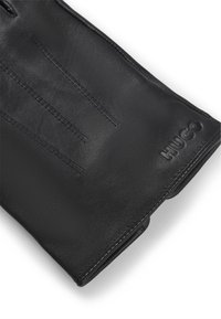 Gant en cuir noir avec une texture lisse, comportant trois lignes cousues au dos et un logo "HUGO" en relief au niveau du poignet.