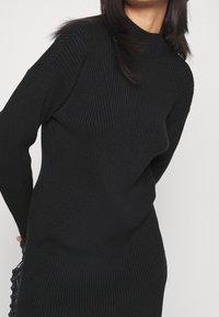 Abito a collo alto nero a costine con maniche lunghe. Presenta un motivo testurizzato e una silhoutte aderente. Realizzato in un morbido materiale lavorato a maglia.