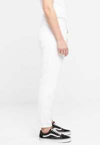 Pantalones de mezclilla blanca con un ajuste slim, piernas cónicas y bolsillos laterales, combinados con zapatillas negras con acentos y cordones blancos.