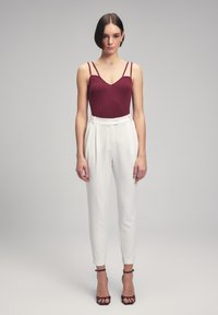 Burgundy ribbed bodysuit with thin straps, paired with high-waisted white pleated trousers. Model wears black strappy heels.
