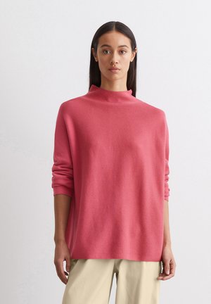 Maglione rosa a maglia con colletto alto e maniche lunghe, caratterizzato da una vestibilità rilassata e tessuto testurizzato. Abbinato a pantaloni di colore chiaro.
