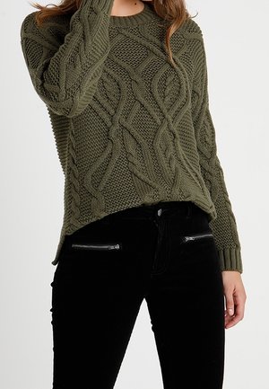 Žena v olivově zeleném svetrům s copánkovým vzorem a černých kalhotách se zipovými kapsami, stojící před jednobarevným pozadím.