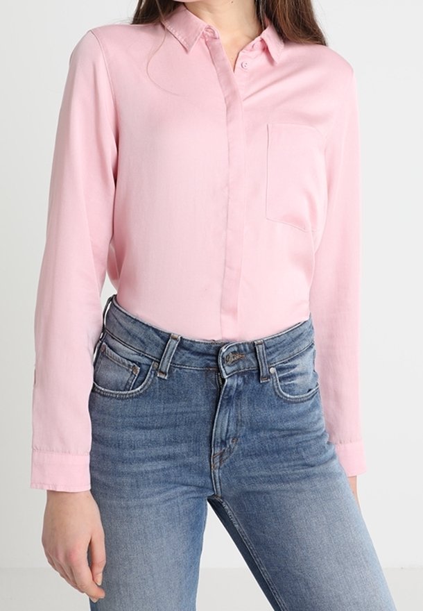Light pink long-sleeve shirt with a button-down collar and chest pocket. Paired with fitted blue denim jeans. Smooth fabric texture.
