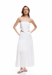 Top blanco sin tirantes con dobladillo en forma de corazón, combinado con una falda larga blanca que presenta patrones de ojal y una cintura fruncida. Sandalias completan el look.
