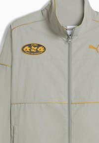 Lys grå lynlåsjakke med gul piping, Puma-logo på højre bryst og Sonic the Hedgehog-patch på venstre bryst.