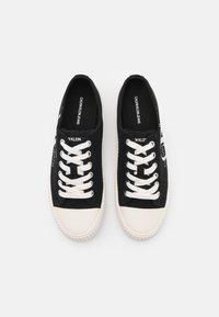 Ett par svarta lågskor i canvas med vita gummikapslar på tårna, vita snören och "Calvin Klein Jeans"-logotyp på innersulor och sidor.