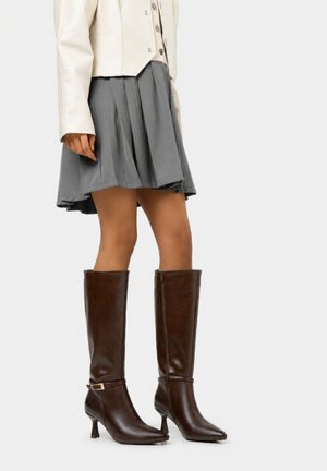 Mujer vistiendo una falda plisada gris, chaqueta crema y botas de cuero marrón oscuro con tacón hasta la rodilla, de pie frente a un fondo liso.