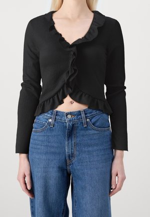 Kobieta w czarnej, długorękawowej, marszczonej, krótkiej bluzce i wysokim stanem niebieskich dżinsów, pokazująca kolczyk w pępku.