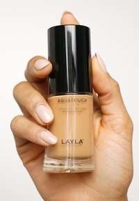 Aquatouch vitamin C-infunderad foundation i en klar glasflaska med svart lock. Slät, krämig textur, medelbeige färg, hand som håller flaskan.