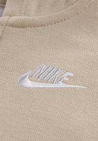 Kreminės spalvos audinys su matoma tekstūra, ant priekinės dalies pavaizduota balta siuvinėta Nike logotipas. Kraštuose yra lygūs siuvimo detalės.