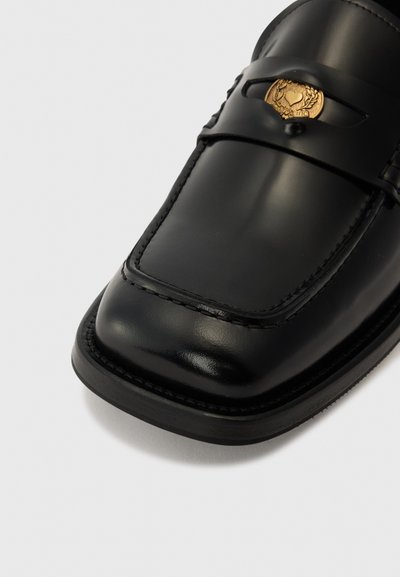 Musthave mustad nahkkingad lamedate ruudukujuliste varvastega, detailse õmblusega ja kuldse embleemi aktsentiga rihm üle ülaosa.
