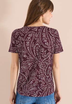 Kvinde iført en kortærmet vinrød top med hvide paisley-blomsterprint, kombineret med blå denimbukser, stående foran en neutral baggrund.
