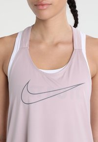 Koszulka na ramiączkach Nike w jasnoróżowym kolorze, wykonana z gładkiego, lekkiego materiału. Posiada szare logo oraz tył w stylu racerback z białymi ramiączkami pod spodem.