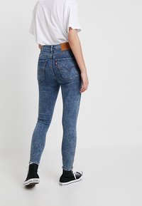 Jeans de tiro alto de color azul claro, con corte ajustado, que cuentan con una etiqueta de cuero en la parte trasera y un diseño clásico de cinco bolsillos. Zapatillas negras.