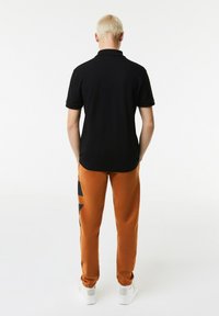 Polo noir à manches courtes, associé à un pantalon de survêtement orange avec un motif graphique noir, porté avec des baskets blanches.