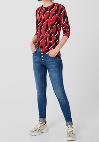 Czerwony longsleeve z czarno-białym abstrakcyjnym wzorem, w połączeniu z dopasowanymi niebieskimi dżinsami z zapięciem na guziki. Jasne sneakersy.
