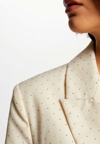 Blazer beige con un tejido texturizado adornado con pequeñas tachuelas plateadas, solapa de pico y detalles de costura visibles en el cuello.