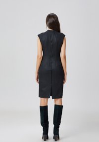 Robe sans manches en suède noir au design ajusté, avec fermeture éclair au dos, ourlet fendu et accents sur les épaules, accompagnée de bottes vertes à mi-mollet.