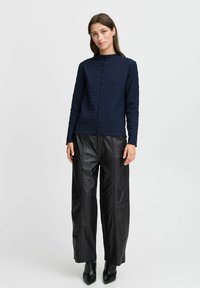 Haut texturé marine avec un col haut, associé à un pantalon en cuir noir ample. La tenue présente un design minimaliste sans motifs visibles.