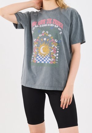 Kvinde iført en grå T-shirt med sol, måne, blomst og ternet mønster, kombineret med sorte cykelshorts.