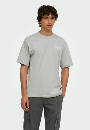 Giovane con capelli ricci che indossa una maglietta grigio chiaro con il logo "modul" e pantaloni cargo grigio scuro, in piedi con una mano in tasca.