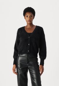 Cardigan nero peloso con scollatura a V, maniche lunghe e bottoni frontali. Abbinato a pantaloni in pelle neri a vita alta. Texture morbida.