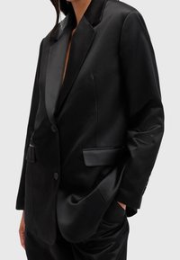 Černý saténový blazer s klopovým límcem, dvěma předními kapsami a zapínáním na jeden knoflík. Hladká textilie s přiléhavým střihem.