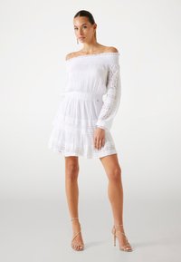 Abito bianco off-the-shoulder con dettagli in pizzo, design a strati e vita elastica. Modella che indossa tacchi dorati con cinghie. Tessuto dalla superficie liscia.