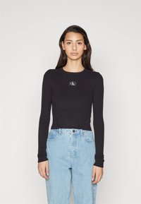 Sort ribbet langærmet top med rund halsudskæring og en rektangulær logo-patch på brystet. Parret med lyse denim jeans i afslappet pasform.